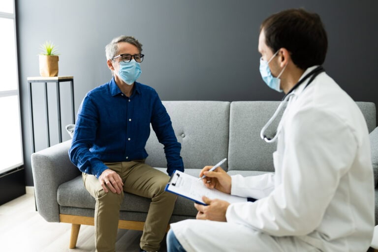 Doctor With Senior Patient Living At Home In Mask